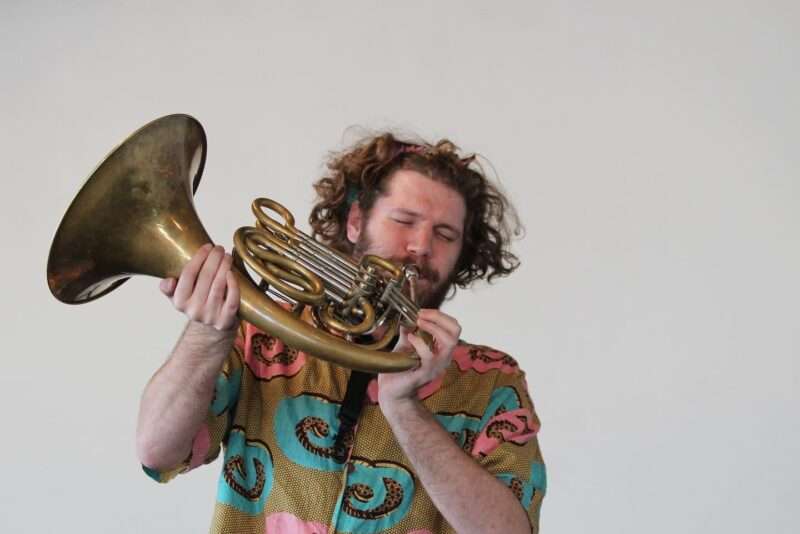 A young man with curly reddish long hair plays a French horn with so much passion that his eyes are closed. He holds the horn in a non-standard way