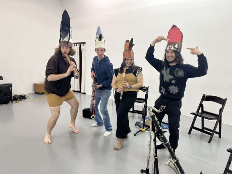Four musicians pose, barefoot or in socks, in a rehearsal space, each wearing a fancy hat that evokes (perhaps pokes fun at) the hat the pope wears. They are all smiling or mugging for the camera.