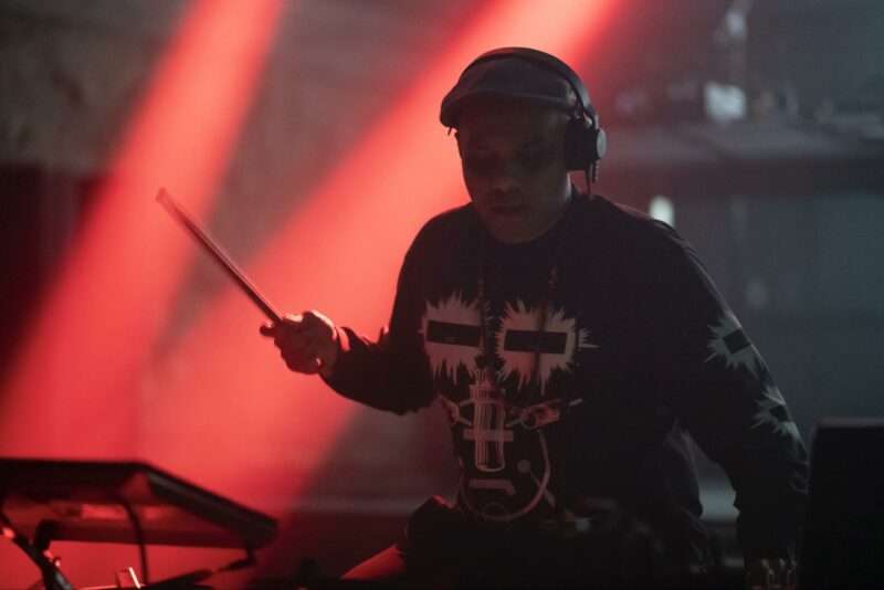 A drummer wearing earphones, a long-sleeve t-shirt with a pattern of black stripes on white, sits at a drum set, one stick in an upraised right hand like he’s mid strike, on a stage in an atmosphere that is dark with bands of pinky red light in back of him.