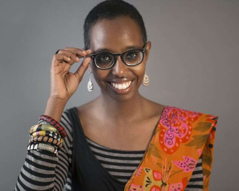 A Black woman with short cropped hair smiles widely and touches her black eyeglasses with her right hand like she’s adjusting them, she is wearing a black and grey striped shirt, colorful bracelets and scarf and prominent white earrings.