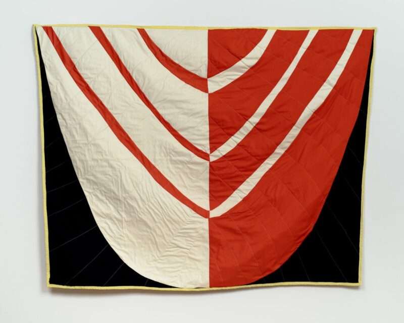 A large square quilted cotton cloth wall hanging with a tongue-like shape dipping down against a black background, with the left half of the tongue showing red stripes on white cloth and the right side showing white stripes on red cloth. The border is yellow.