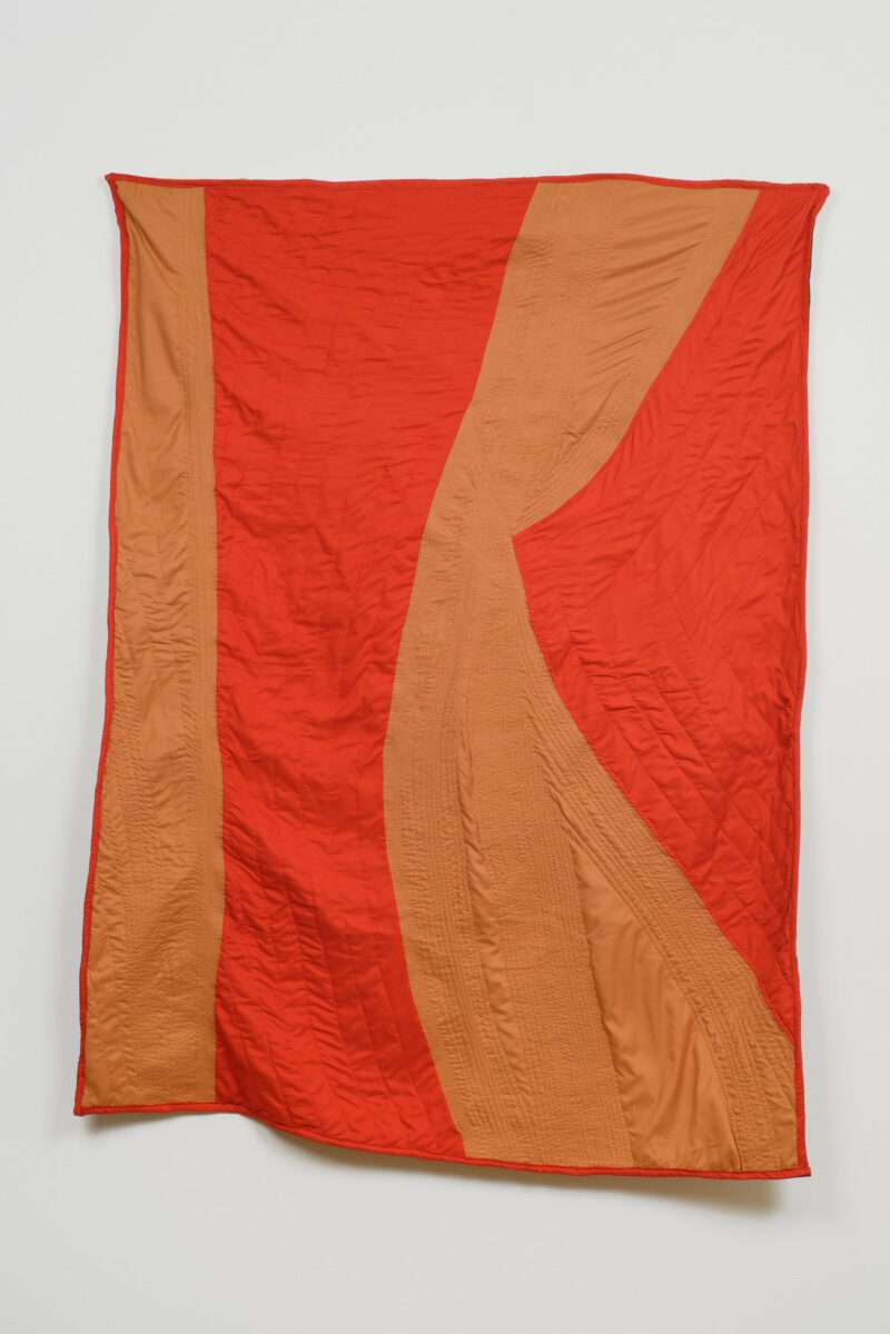 A large quilted cotton cloth wall hanging has orange and red colored fields alternating in uneven strips, with the sewing displayed prominently, almost like rows of a farm field that follow the lay of the land.