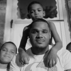 A black and white portrait photograph shows a man and three boys gathered together and smiling while sitting on a stoop in front of their front door.