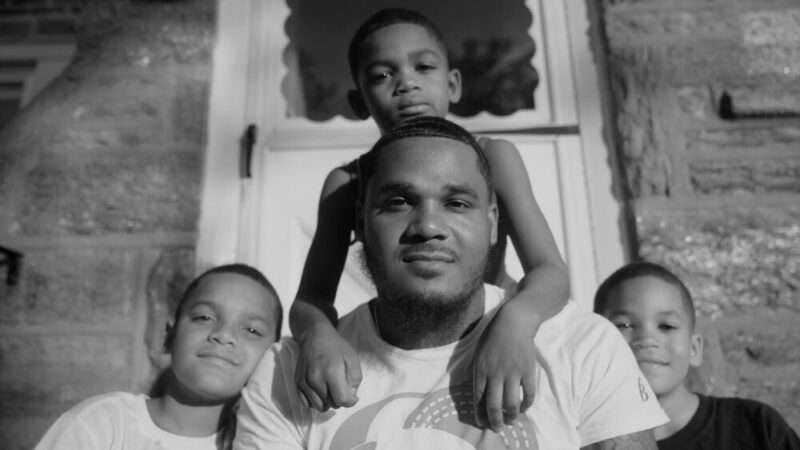 A black and white portrait photograph shows a man and three boys gathered together and smiling while sitting on a stoop in front of their front door. 