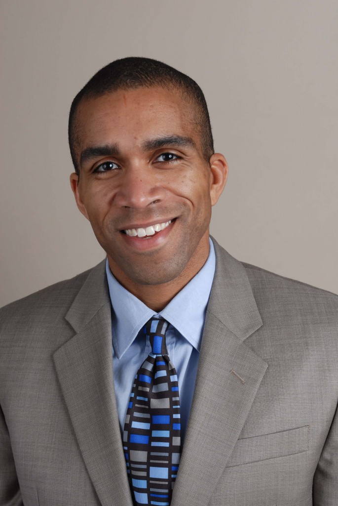 photo of the head and shoulders of a smiling African American man, Blake Bradford, in a gray suit, blue shirt and patterned tie.d