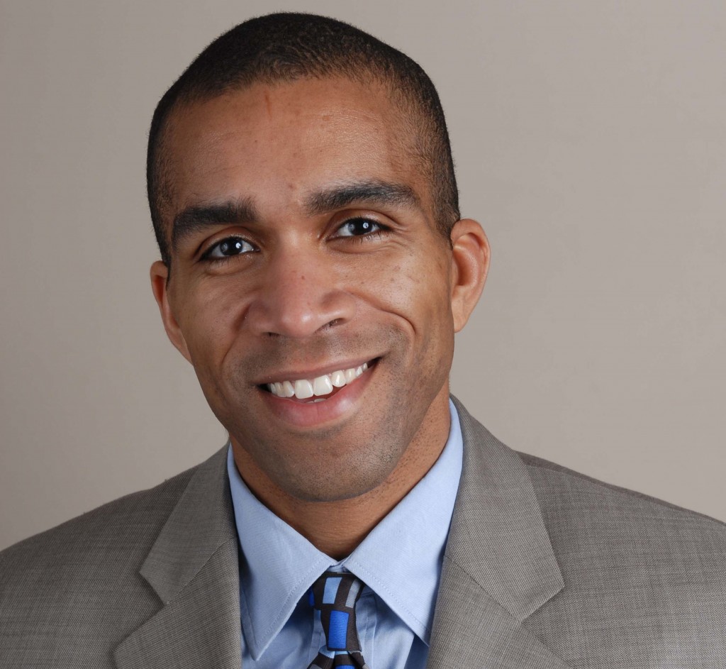 Blake Bradford, a Black man with buzzed dark hair, wearing a gray suit, blue shirt, and patterned tie, and is smiling.