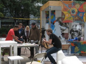 Pedro Ospina (left) drawing with passersby, klip collective taping. A charcoal portrait by Betsy Casanas hangs on the Welcome House.